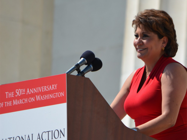 Color photo of smiling woman standing at lectern with microphone and sign printed on front, reading, “The 50th Anniversary of the March on Washington: National Action to Realize the Dream.” 